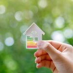 A Hand Holds a Small Paper House With Bars Colored From Green to Red Across It.