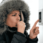 A person wearing a winter coat and looking cold, adjusts their thermostat.