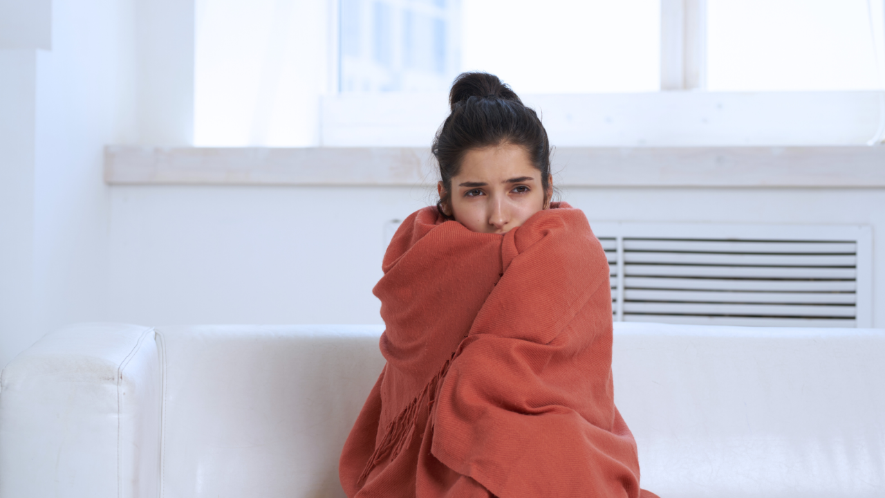 A woman wraps a blanket around herself to protect from cold in her home.