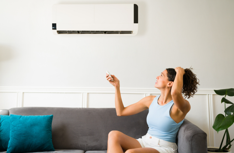 A woman in a light blue top sits on a gray couch with teal pillows, pointing a remote at a white wall-mounted AC unit.