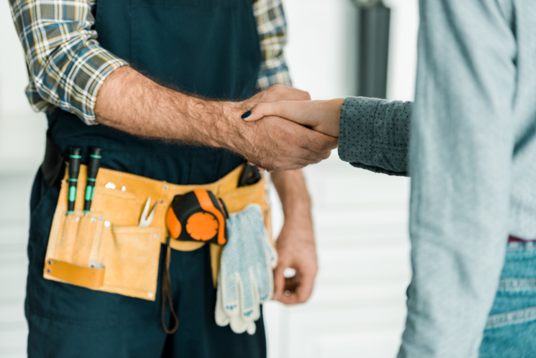 HVAC technician in navy apron and plaid shirt shakes hands with homeowner in gray sweater while wearing a yellow leather tool belt.